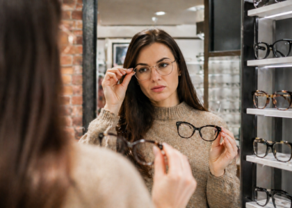Kobieta przymierza damskie okulary przed lustrem w salonie optycznym i porównuje dwa modele oprawek.